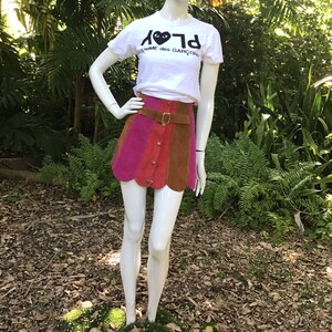 May include: A white t-shirt featuring a black heart with eyes graphic and the text "PLAY" in reverse. The shirt is worn with a multi-coloured suede mini skirt and a brown leather belt.