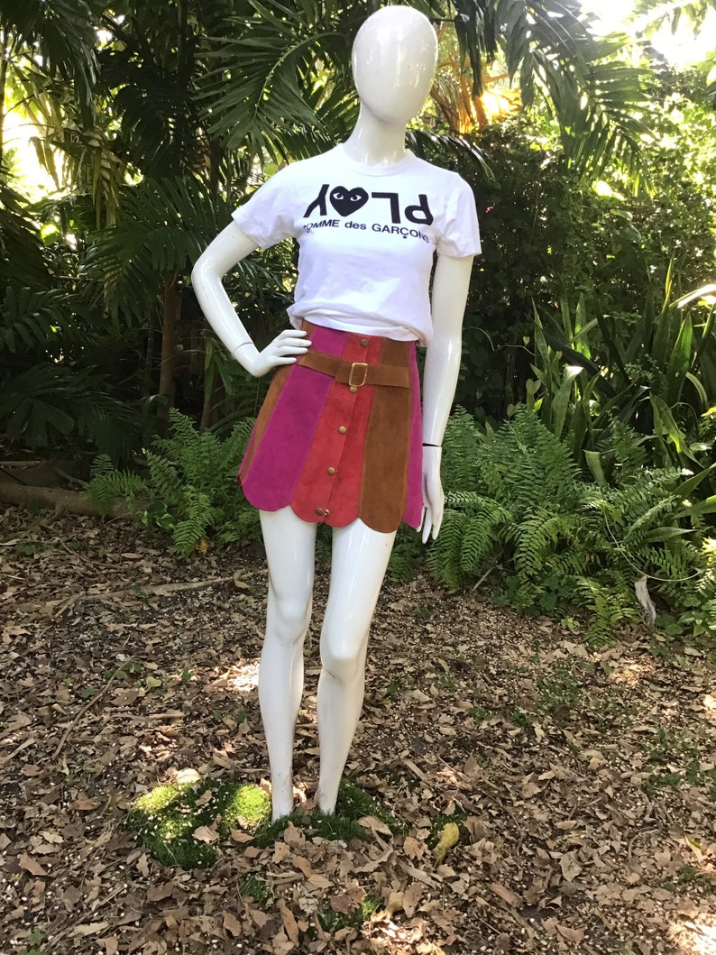 May include: A white t-shirt featuring a black heart with eyes graphic and the text "PLAY" in reverse. The shirt is worn with a multi-coloured suede mini skirt and a brown leather belt.