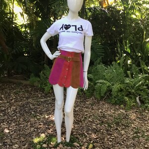 May include: A white t-shirt with a black heart and the text "PLAY" and "COMME des GARCONS" on it. The person is wearing a multi-coloured mini skirt with a brown leather belt.