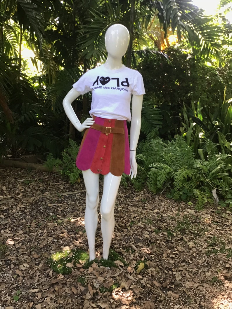 May include: A white t-shirt with a black heart and the text "PLAY" and "COMME des GARCONS" on it. The person is wearing a multi-coloured mini skirt with a brown leather belt.