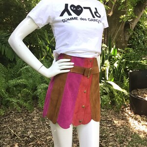 May include: A brown, pink and red suede mini skirt with a brown leather belt and button fastening. The skirt has a scalloped hem and is being worn by a mannequin. The mannequin is wearing a white t-shirt with the text "PLAY COMME des GARCONS" printed on it.