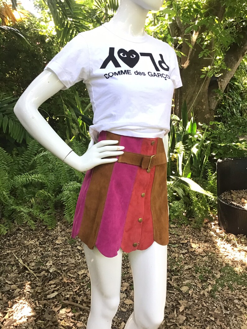 May include: A brown, pink and red suede mini skirt with a brown leather belt and button fastening. The skirt has a scalloped hem and is being worn by a mannequin. The mannequin is wearing a white t-shirt with the text "PLAY COMME des GARCONS" printed on it.