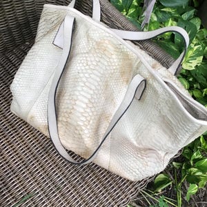 May include: A white leather tote bag with a snakeskin pattern. The bag has two top handles and a zipper closure.