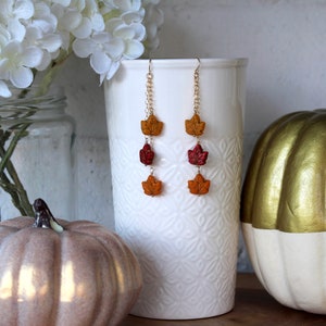 Op de afbeelding: Een paar hangende oorbellen met drie herfstkleurige bladeren aan elke oorbel. De bladeren zijn in tinten oranje, rood en geel. De oorbellen hangen aan een gouden ketting.