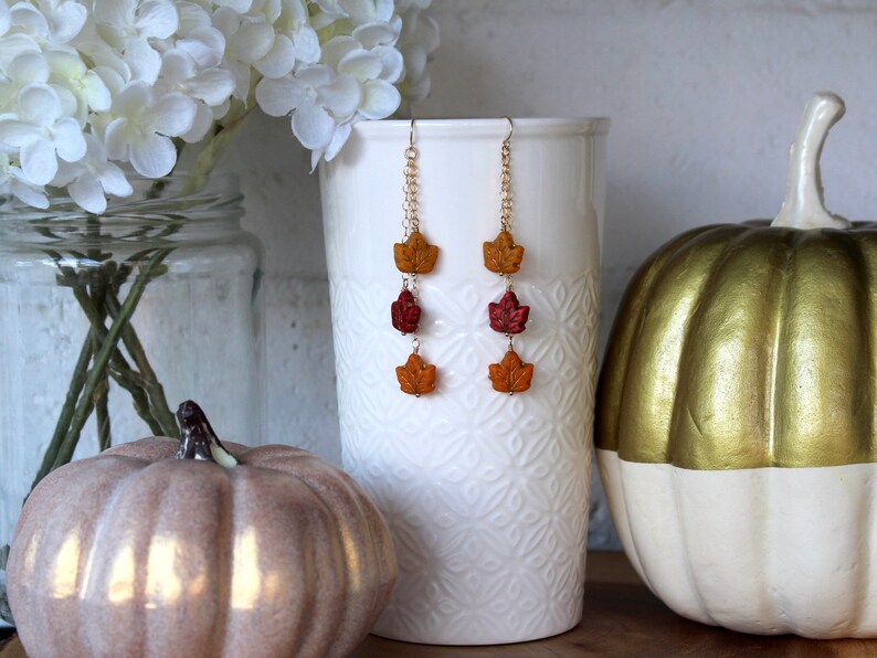 Op de afbeelding: Een paar hangende oorbellen met drie herfstkleurige bladeren aan elke oorbel. De bladeren zijn in tinten oranje, rood en geel. De oorbellen hangen aan een gouden ketting.