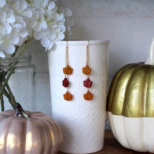 Op de afbeelding: Een paar lange hangende oorbellen met drie bladeren in herfstkleuren aan elke oorbel. De bladeren zijn oranje, rood en geel. De oorbellen hangen aan een gouden ketting.