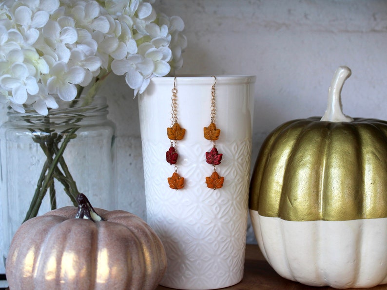 Op de afbeelding: Een paar lange hangende oorbellen met drie bladeren in herfstkleuren aan elke oorbel. De bladeren zijn oranje, rood en geel. De oorbellen hangen aan een gouden ketting.