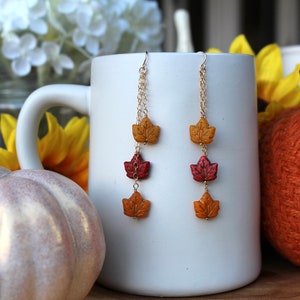 Op de afbeelding: Een paar hangende oorbellen met drie herfstkleurige bladeren aan elke oorbel. De bladeren zijn gemaakt van polymer clay en zijn geschilderd in tinten rood, oranje en geel. De oorbellen hangen aan gouden kettingen.