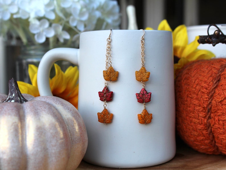 Op de afbeelding: Een paar hangende oorbellen met drie herfstkleurige bladeren aan elke oorbel. De bladeren zijn gemaakt van polymer clay en zijn geschilderd in tinten rood, oranje en geel. De oorbellen hangen aan gouden kettingen.