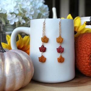 Op de afbeelding: Een paar gouden oorhangers met drie blaadjesvormige kralen in tinten geel, oranje en rood. De oorhangers hangen aan een witte mok.