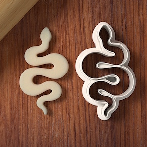Puede incluir: Un cortador de galletas de plástico blanco en forma de serpiente sobre una superficie de madera. Una galleta en forma de serpiente blanca está al lado del cortador.