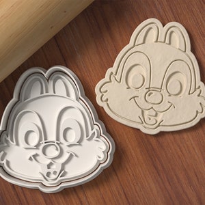 May include: Two white plastic cookie cutters shaped like the face of a cartoon chipmunk. The cookie cutters are on a wooden surface.
