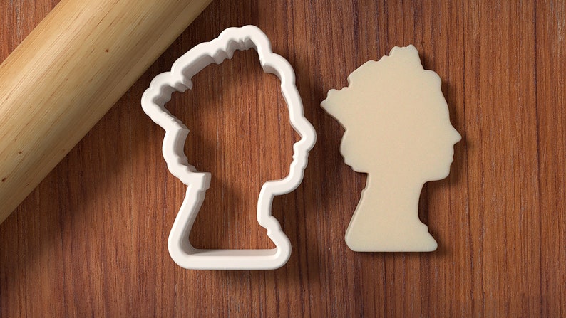 May include: A white plastic cookie cutter shaped like a profile of a person wearing a crown. The cutter is on a wooden surface next to a rolling pin.