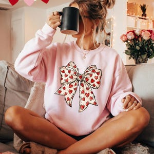 May include: A light pink sweatshirt featuring a large bow design with red hearts. The bow has a cream-colored background with red heart accents. The sweatshirt is a crew neck style and is made of a soft material.