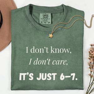May include: A sage green t-shirt with the text "I don't know, I don't care, IT'S JUST 6-7." A gold star pendant necklace rests on the shirt. The shirt is folded, and a brown hat is visible in the background.