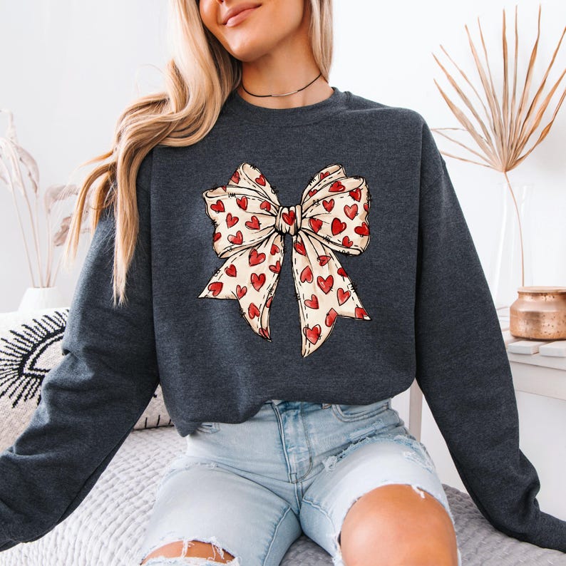 May include: Dark gray sweatshirt featuring a large bow design. The bow is cream-colored with a pattern of red hearts. The sweatshirt is a casual top, suitable for everyday wear.