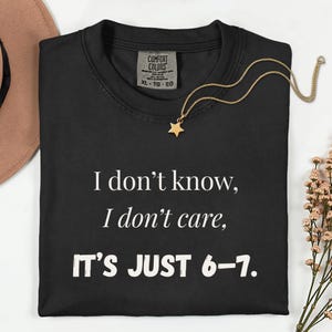 May include: Black t-shirt with the text "I don't know, I don't care, IT'S JUST 6-7." A gold star pendant necklace rests on the shirt. The shirt is labeled XL-TG-EG.