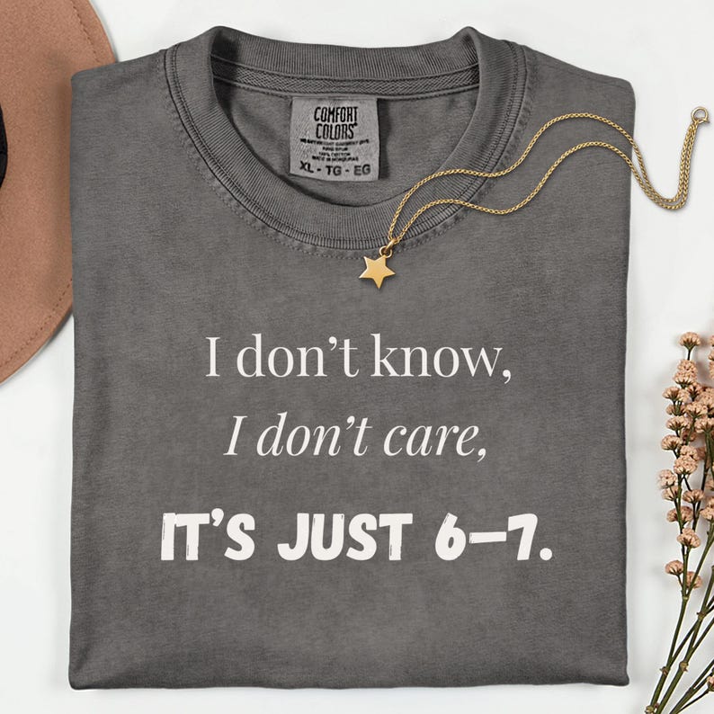 May include: A dark gray Comfort Colors t-shirt with the text "I don't know, I don't care, IT'S JUST 6-7." A gold star pendant necklace rests on the shirt. The shirt is XL size.
