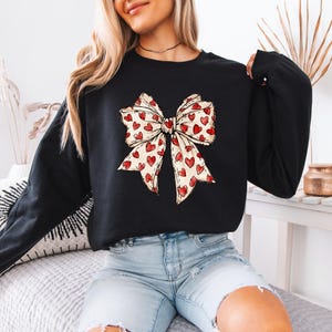 May include: Black sweatshirt featuring a large bow design. The bow is cream-colored with a pattern of red hearts. The sweatshirt has long sleeves and a crew neck. The overall style is casual and suitable for Valentine's Day or everyday wear.