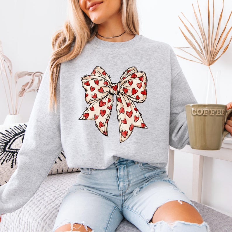 May include: A light gray sweatshirt featuring a large bow design. The bow is cream-colored with a pattern of small red hearts. The sweatshirt is a casual style, suitable for everyday wear.