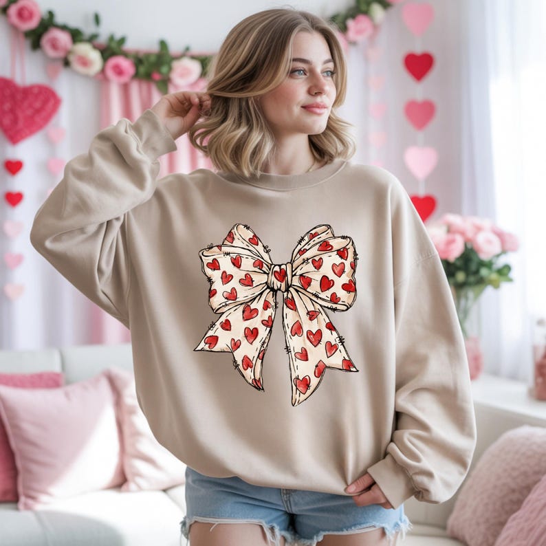 May include: A beige sweatshirt featuring a large bow design. The bow is cream-colored with red heart patterns. The sweatshirt is a neutral color, suitable for casual wear. The design is centered on the front of the garment.