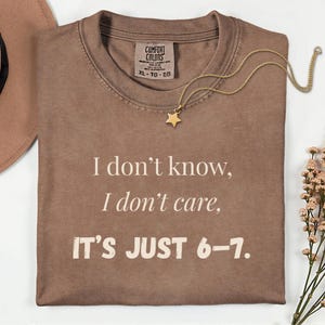 May include: A brown Comfort Colors t-shirt with the text "I don't know, I don't care, IT'S JUST 6-7." A gold star pendant necklace rests on the shirt. The shirt is folded and lies on a white surface.