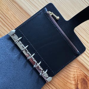 May include: Black leather binder with silver metal rings and a zippered pocket. The binder is open, revealing the interior pocket with a gold-colored zipper pull. The leather has a smooth texture, and the binder is set against a wooden surface.