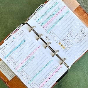 May include: A personal planner with a brown leather cover and a six-ring metal binder mechanism. The white paper pages feature printed monthly calendars, lined sections for daily notes, and handwritten entries in blue, pink, and black ink.
