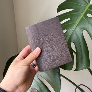 May include: A small, textured, brown leather wallet. The wallet is rectangular and appears to be closed. The leather has a pebbled texture. The background includes a large green plant with large leaves.