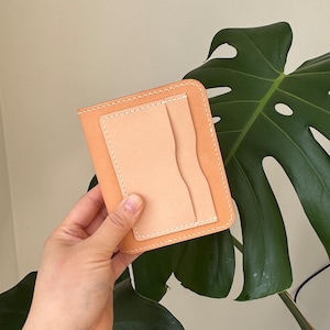 May include: A light brown leather wallet with a lighter tan interior and a stitched border. The wallet has a rectangular shape with rounded corners and a vertical pocket on the front.