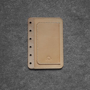 May include: A tan leather notebook cover with a small pocket on the front. The cover has a dog face logo and six holes on the left side for a binder.