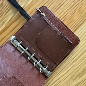 May include: A brown leather personal organizer featuring a six-ring silver metal binder mechanism, a zippered pocket with a small gold-toned pull, and a smooth leather interior pocket. A black elastic band is attached to the top edge of the cover.