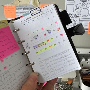 May include: A planner open to a March 2025 calendar page, with highlighted dates and handwritten notes. The planner has a pink sticky note and a tab labelled "3月 스케줄". The planner is held open with a black strap.