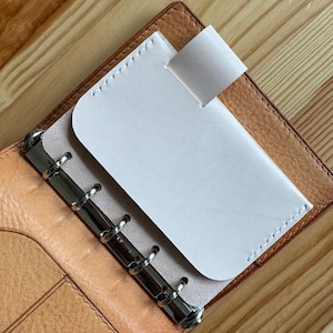 May include: Open leather planner with a light tan interior and a brown exterior. A white card holder is inserted into the planner, held in place by a white tab. The planner has silver metal rings.