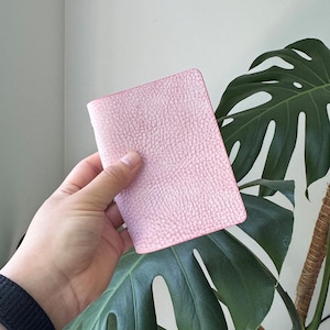 May include: A pink, textured leather notebook with a matching pink border. The notebook is held in a hand, with a large green plant in the background.