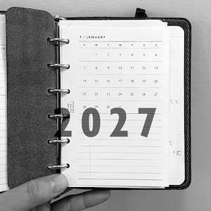 May include: A black ring-bound planner open to a January calendar page. The page displays the text 1 / JANUARY, a grid of dates from 1 to 31, and the text Pocket THINK THANK AT. The large bold text 2027 is overlaid across the centre of the page.