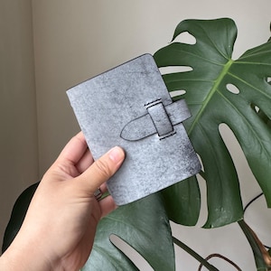 May include: A grey leather journal with a buckle fastening. The journal has a textured surface and is rectangular. The buckle is also grey and appears to be made of the same material. The journal is held in front of a large green leaf.