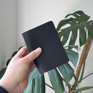 May include: A black, textured leather notebook held in a hand. The notebook is rectangular with rounded corners. The background includes large green leaves of a houseplant and a white wall.