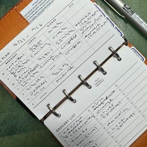 May include: A ring-bound planner insert with a tan leather cover, featuring a weekly meal plan grid and a grocery list section. The white paper contains handwritten entries for breakfast, lunch, and dinner, along with a list of items with circular checkboxes.