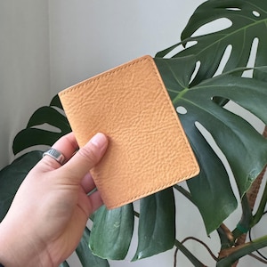 May include: A tan leather wallet with a textured surface. The wallet is rectangular and appears to be hand-stitched with a contrasting thread. The wallet is held in a hand, with a silver ring visible on the finger.