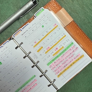 May include: An open planner showing a May calendar with highlighted dates and handwritten notes. A silver pen lies next to the planner, which is in a brown leather cover. The planner has tabs and a metal ring binder.
