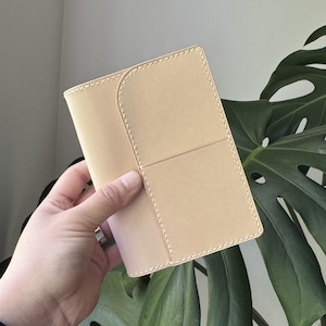 May include: A light beige leather wallet with a curved flap and stitched detailing. The wallet has a rectangular shape with a divided front panel. The stitching is a light cream color. The wallet is held in a person's hand.