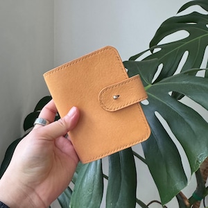 May include: A tan leather wallet with a flap closure and a silver-colored snap. The wallet is held in a hand, with a silver ring on the finger. The background features large green leaves.