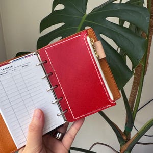 May include: Red leather planner with white stitching and a zipper pocket. The planner is open, revealing lined pages for notes and a tracker. The planner is held open with silver rings.