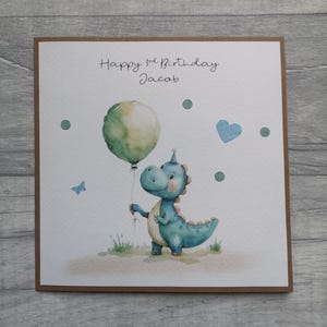 May include: A birthday card featuring a watercolor illustration of a blue dinosaur holding a green balloon. The card reads "Happy 3rd Birthday Jacob" in handwritten script, with additional decorative elements like a blue heart and a butterfly.