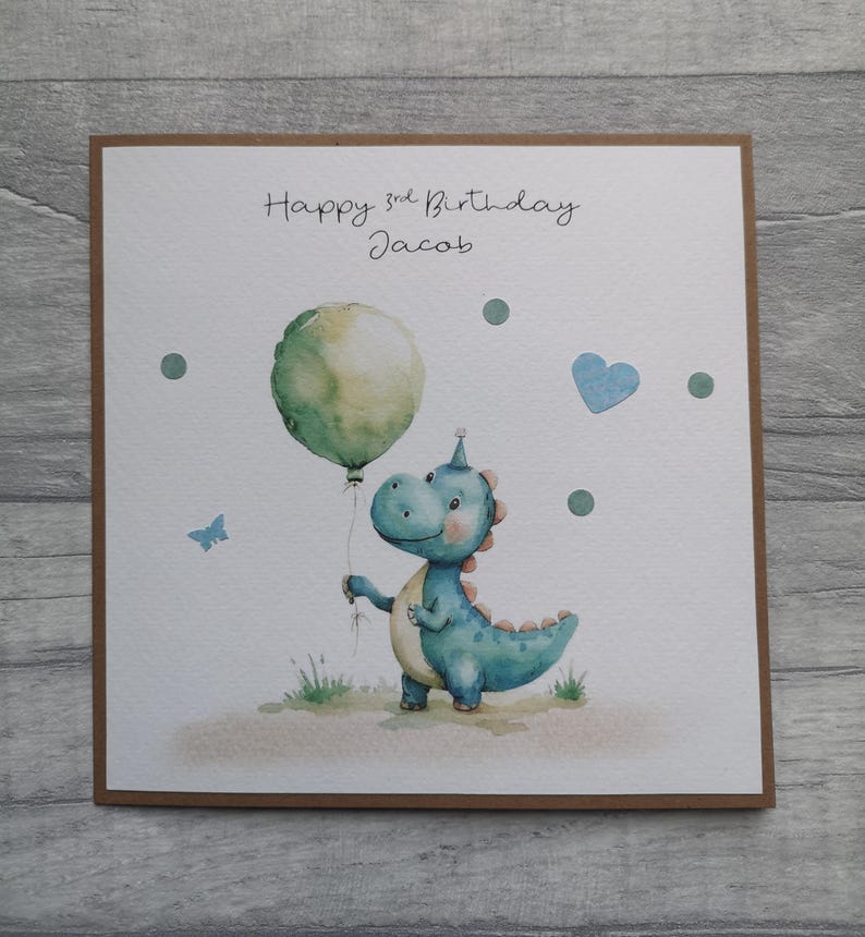 May include: A birthday card featuring a watercolor illustration of a blue dinosaur holding a green balloon. The card reads "Happy 3rd Birthday Jacob" in handwritten script, with additional decorative elements like a blue heart and a butterfly.