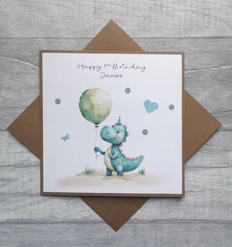 May include: A watercolor birthday card featuring a blue dinosaur holding a green balloon. The card reads "Happy 3rd Birthday Jacob" in black script. Additional details include a blue heart, a butterfly, and green dots.