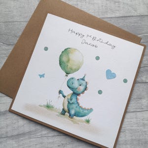 May include: A birthday card featuring a watercolor illustration of a blue dinosaur holding a green balloon. The card reads "Happy 3rd Birthday Jacob" in handwritten script. Additional details include a blue heart, a butterfly, and green dots.