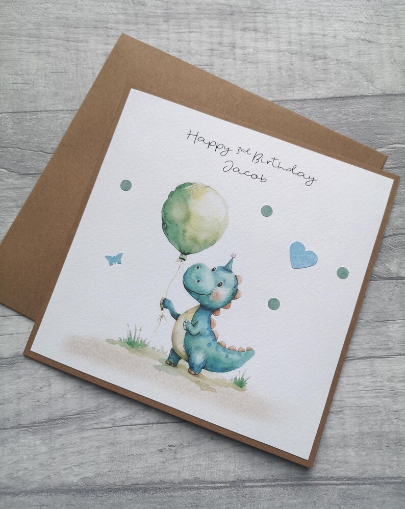 May include: A birthday card featuring a watercolor illustration of a blue dinosaur holding a green balloon. The card reads "Happy 3rd Birthday Jacob" in handwritten script. Additional details include a blue heart, a butterfly, and green dots.