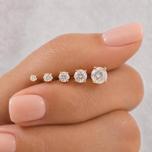 May include: A set of five gold stud earrings, each featuring a single round brilliant-cut clear gemstone held in a four-prong setting. The earrings are arranged in a row by size, from the smallest stone on the left to the largest stone on the right.